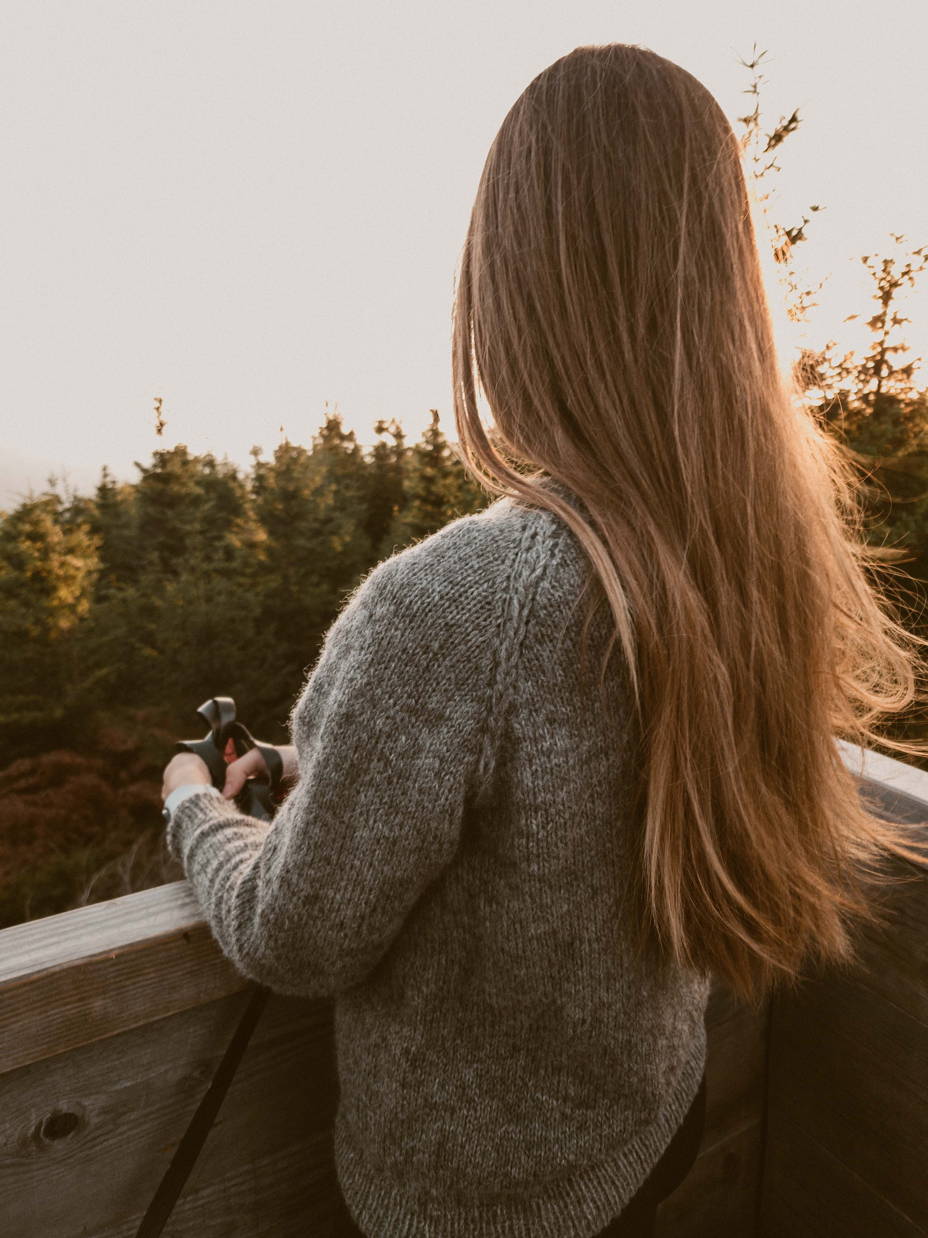 Designer Ulven in a grey Wullpullover sweater handknit in Pommernglück from Calling Sheep, standing on a wooden platform at golden hour with blurred trees in the background.