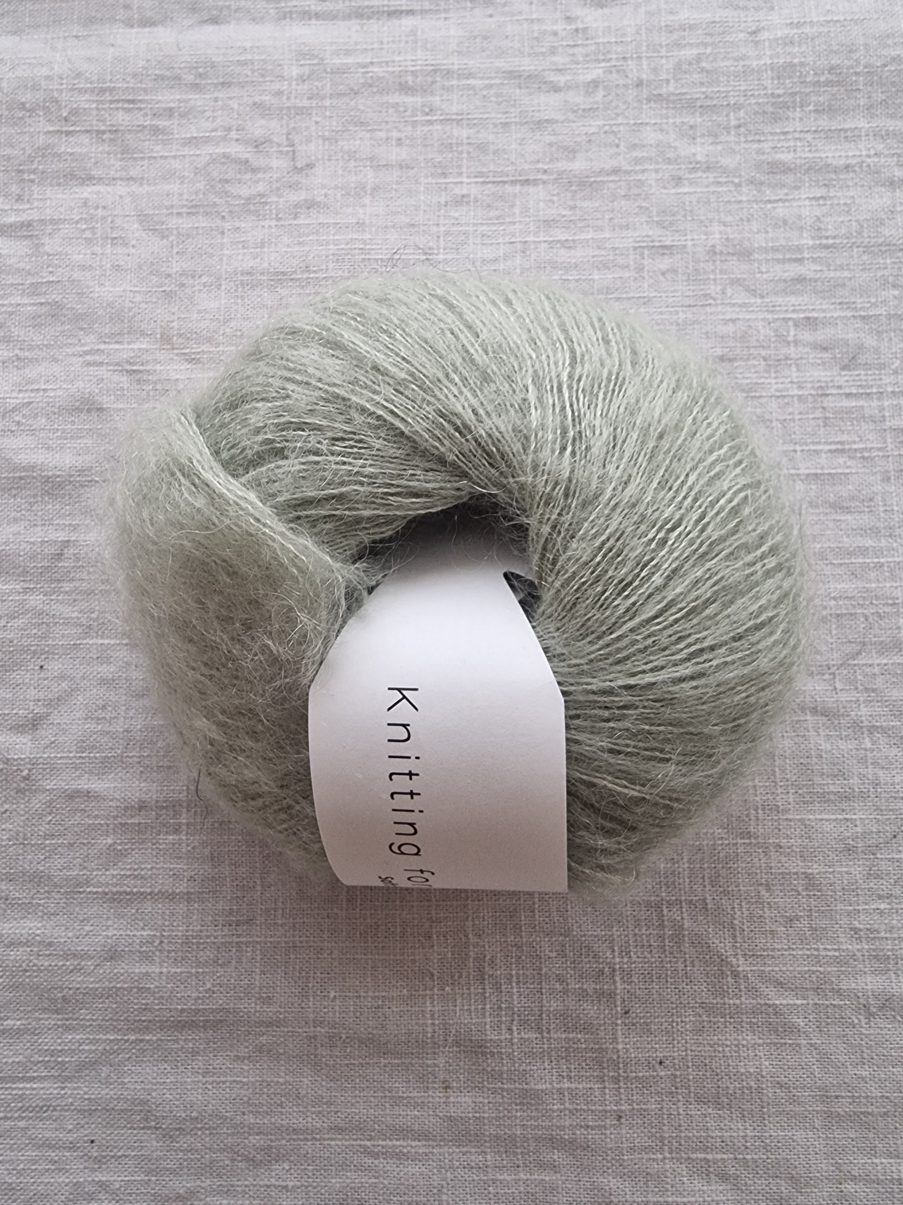 Soft sage green lace weight silk mohair yarn skein with fluffy texture on linen background.