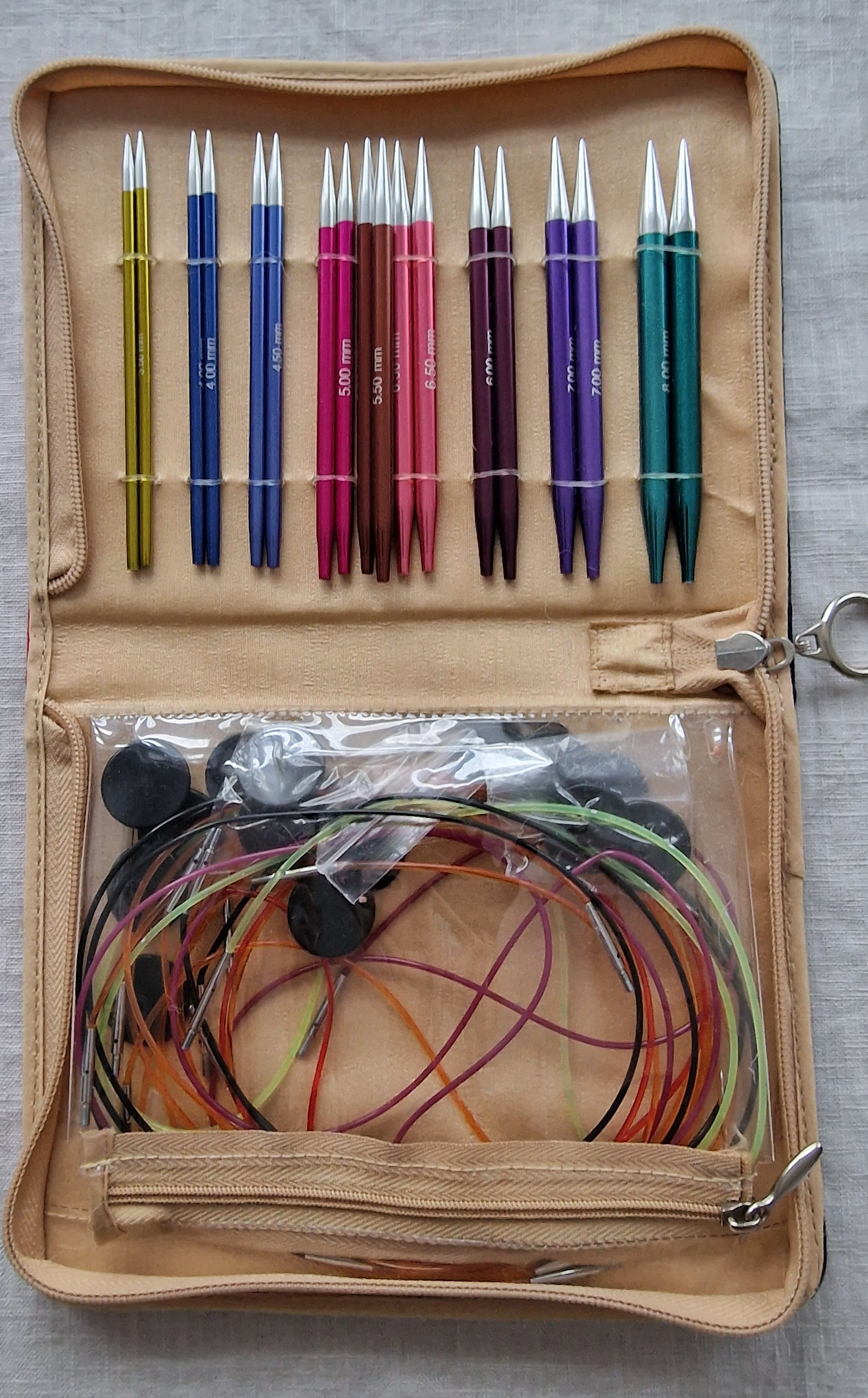 Colorful interchangeable knitting needle set in tan case with multiple cable cords and accessories.