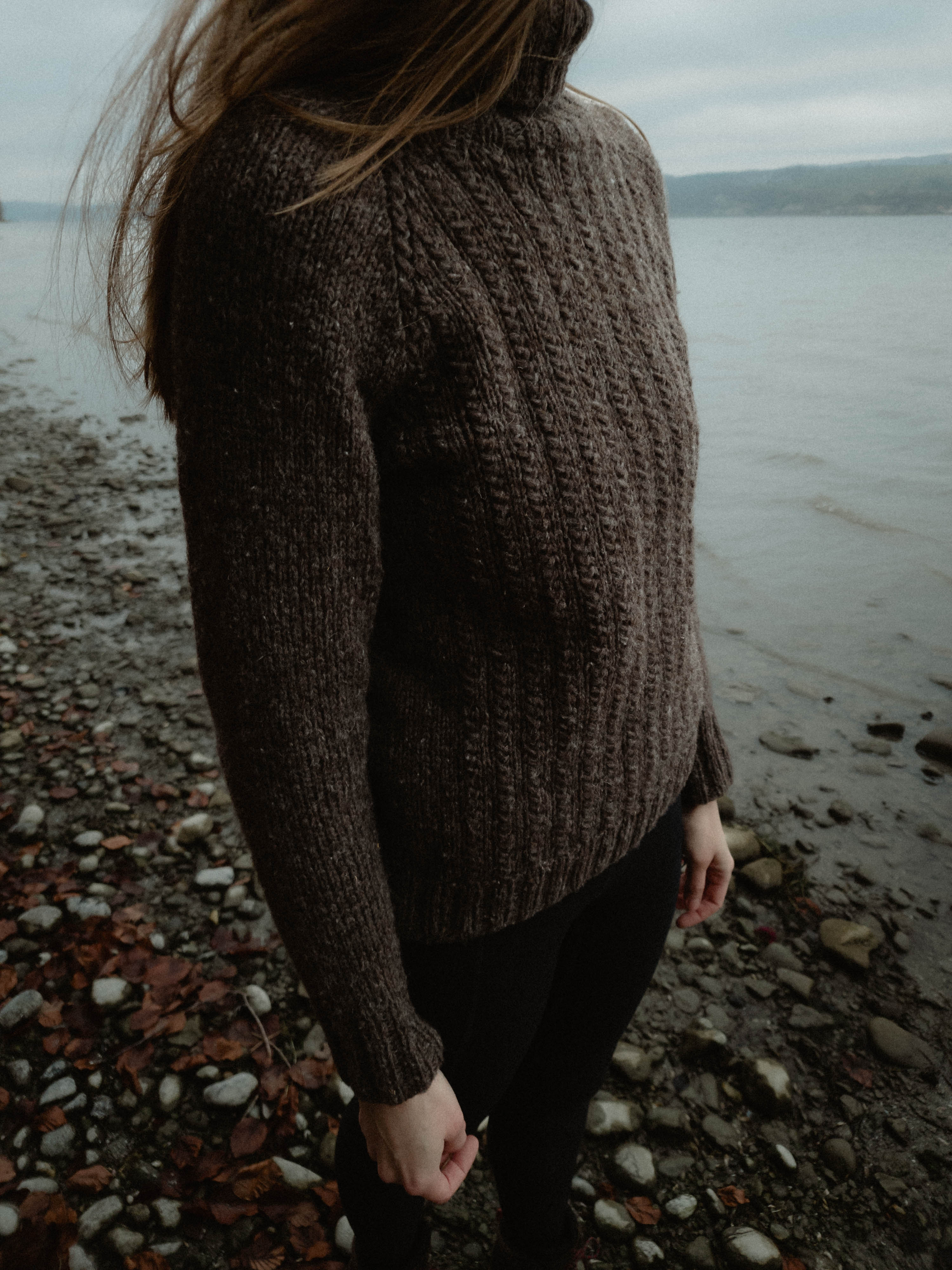 Woman wearing charcoal grey cable knit sweater standing on rocky beach by water.