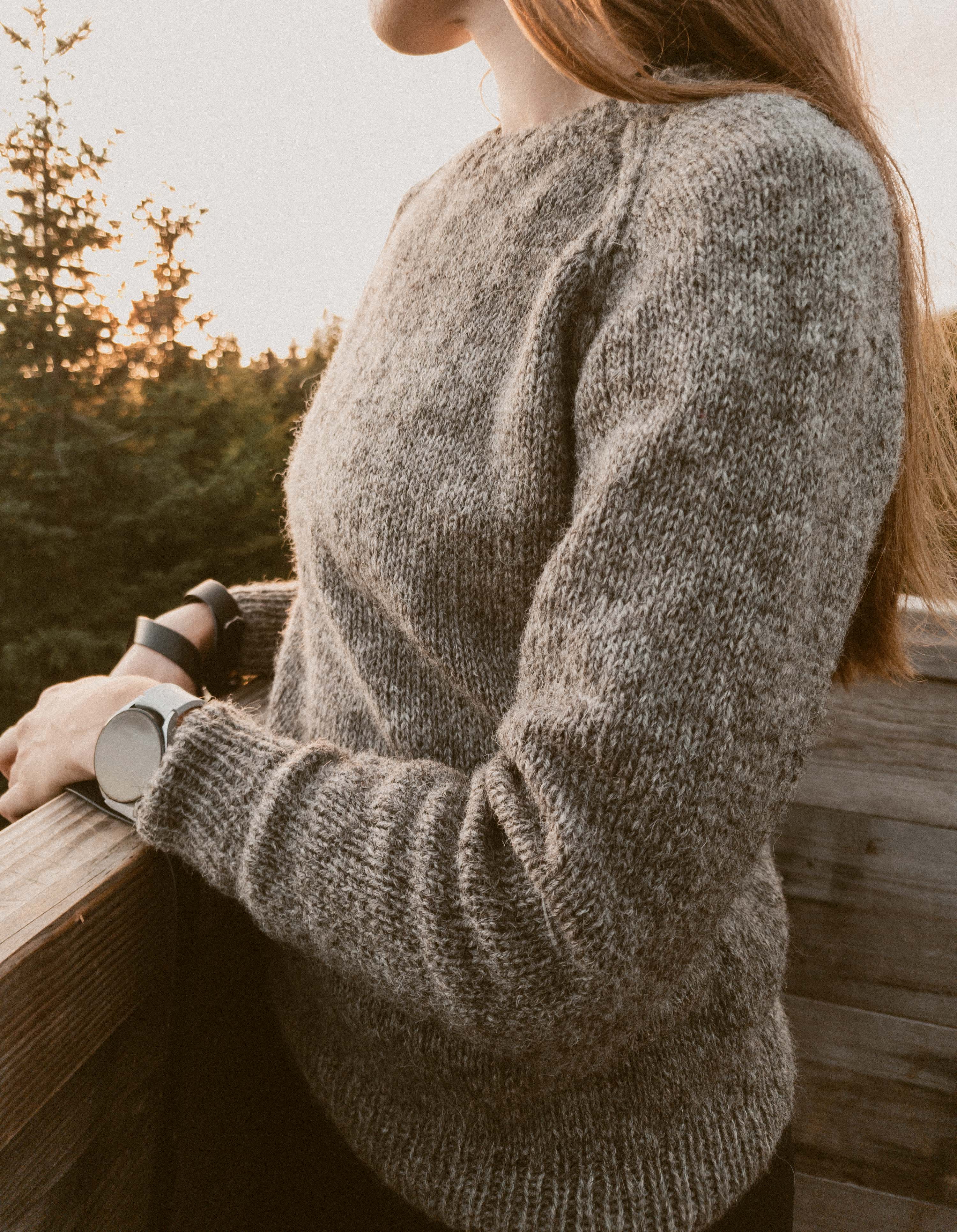 Designer Ulven in a grey Wullpullover sweater handknit in Pommernglück from Calling Sheep, standing on a wooden platform at golden hour with blurred trees in the background.