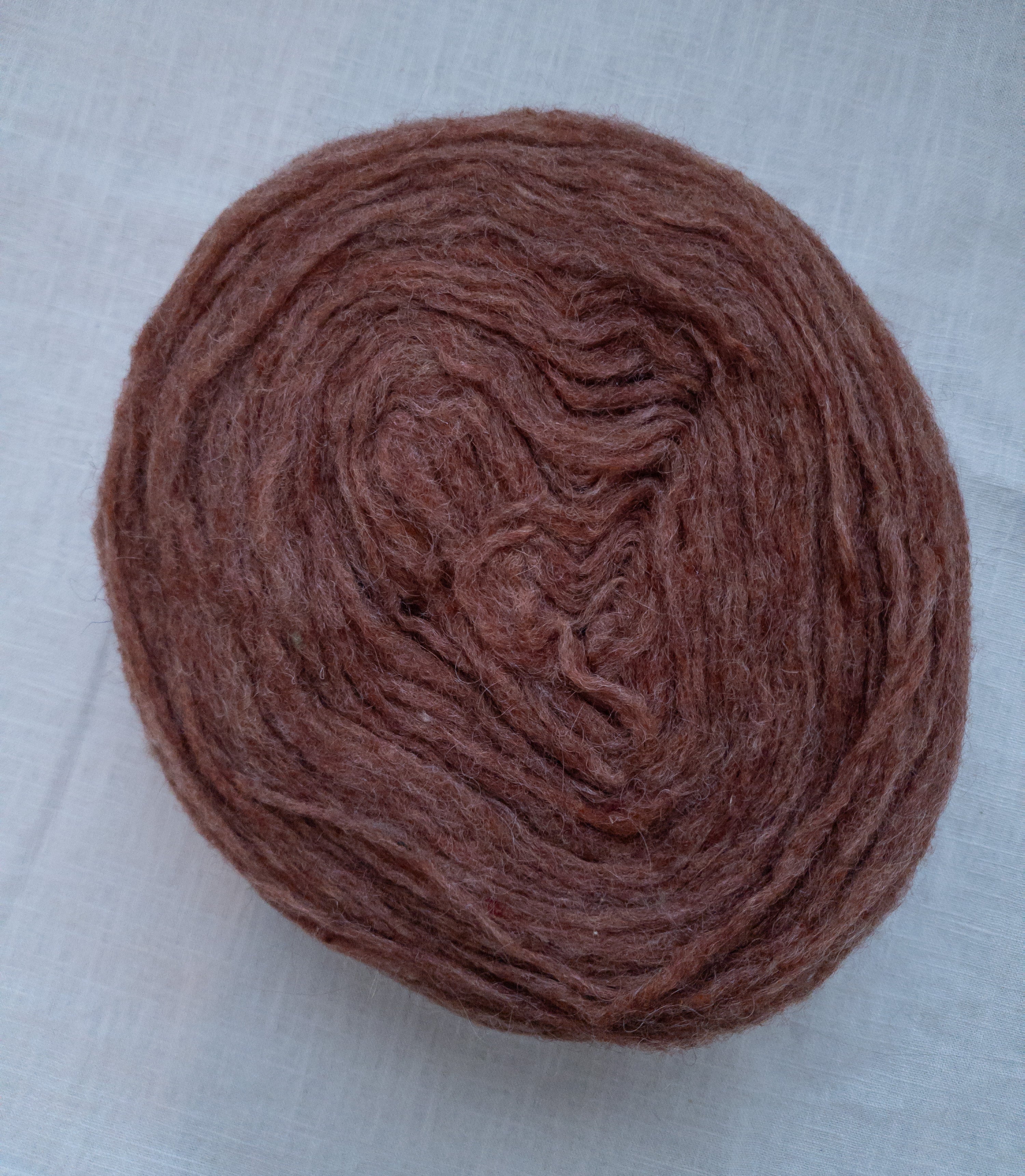 A round cake of unspun brick red Swedish wool yarn with soft fuzzy texture on light fabric background.